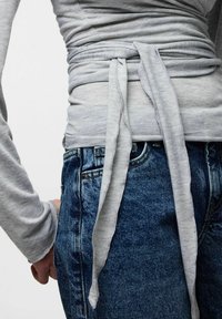 Sivi zgornji del z dolgimi rokavi, ki se zavezuje okoli pasu, v kombinaciji z modrimi visokim pasom kavbojkami iz jeansa. Tekstura je videti mehka in oprijeta.