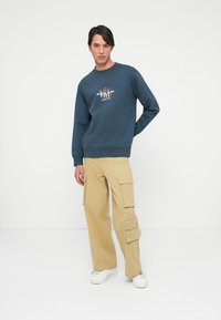 Marineblauwe sweatshirt met ronde hals en grafisch ontwerp op de borst, gecombineerd met beige cargobroek met meerdere zakken. Witte sneakers.