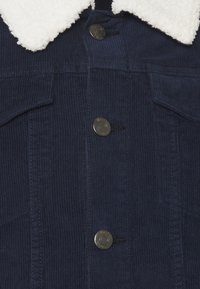 Giacca di velluto a coste blu navy con colletto in pile bianco e tre bottoni metallici sul davanti.