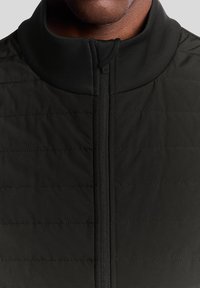 Veste isolante noire avec un col haut, dotée d'une fermeture éclair à l'avant et d'un motif matelassé horizontal sur la poitrine. Texture lisse.