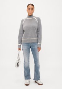 Γκρι πουλόβερ με ψηλό λαιμό, ιβουάρ λεπτομέρειες και ριμπ υφή, συνδυασμένο με μπλε καμπάνα τζιν και μεταλλικά ασημένια παπούτσια, κρατώντας μια ασημένια τσάντα.