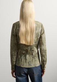 Camicia a maniche lunghe verde oliva con un tessuto strutturato e sgualcito e sottili motivi più scuri, abbinata a jeans blu a vita alta.