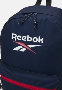 Námornícko modrý batoh Reebok s červeným zipsom, výrazným logom v bielej a červenej farbe, vyrobený z odolného materiálu a s nastaviteľnými popruhmi.