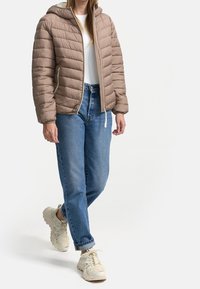 Puffjacka i ljusbrunt, med huva, quiltad textur, framdragkedja och sidofickor. I kombination med blå jeans och beige sneakers.