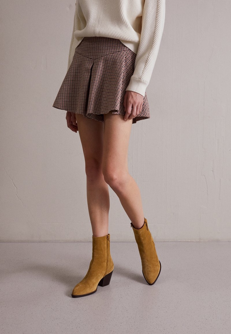 Bruin geruite minirok met plooien, gecombineerd met mosterdsuède enkellaarsjes met een spitse neus en blokhak. Bovenkant een witte gebreide trui.