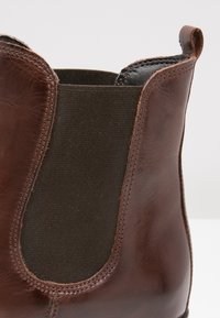 Bottine en cuir marron avec un panneau latéral élastique foncé, un bout rond et des détails cousus. La texture est lisse avec une finition polie.