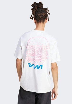 Mand med dreadlocks iført en hvid t-shirt med en pink burgergrafik og blåt krøllet design på bagsiden mod en ensfarvet baggrund.