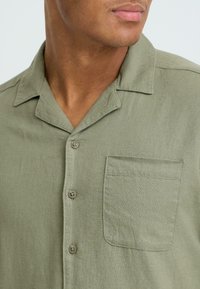 Chemise à manches courtes de couleur vert olive, avec boutons, en tissu texturé. Elle comprend un col, une poche avant et deux boutons marron sur la patte de boutonnage.