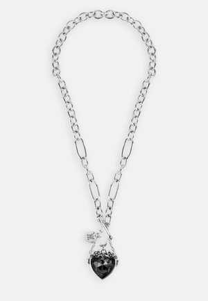 Colar de corrente prateada com um grande pendente em forma de coração negro, adornado com detalhes decorativos e uma pequena etiqueta.