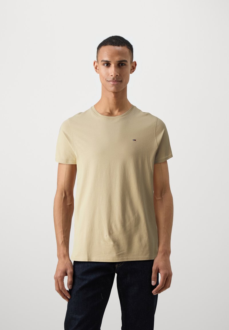Beige pamut póló kerek nyakkivágással és rövid ujjakkal, elöl egy kis logóval. Sötét farmernadrággal párosítva.