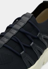 Κοντινό πλάνο μαύρου δίχτυνου αθλητικού παπουτσιού με ελαστικά κορδόνια και δερμάτινους ιμάντες για τα κορδόνια σε λευκή σόλα.