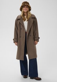 Smešani kaput od smeđeg vune s dvostrukim kopčanjem, širokim reverima i velikim džepovima. Model nosi plave široke traperice i smeđi šešir s uzorkom leoparda.