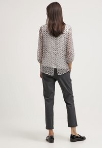Durchsichtige, gemusterte Bluse in hellbeige mit Dreiviertelärmeln, über einem weißen Top getragen. Kombiniert mit dunkelgrauen, hochgekrempelten Hosen und schwarzen Schuhen.