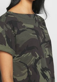 Haut à manches courtes imprimé camouflage dans des tons de vert et de noir, fabriqué en coton doux. Il présente un col large et des manches retroussées.