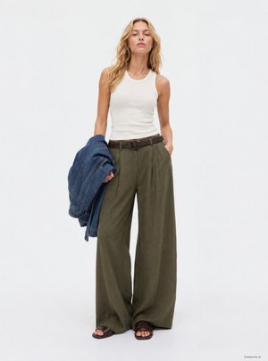 Donna con top bianco senza maniche e pantaloni larghi verde oliva tiene una giacca di jeans in una mano e l'altra mano in tasca.