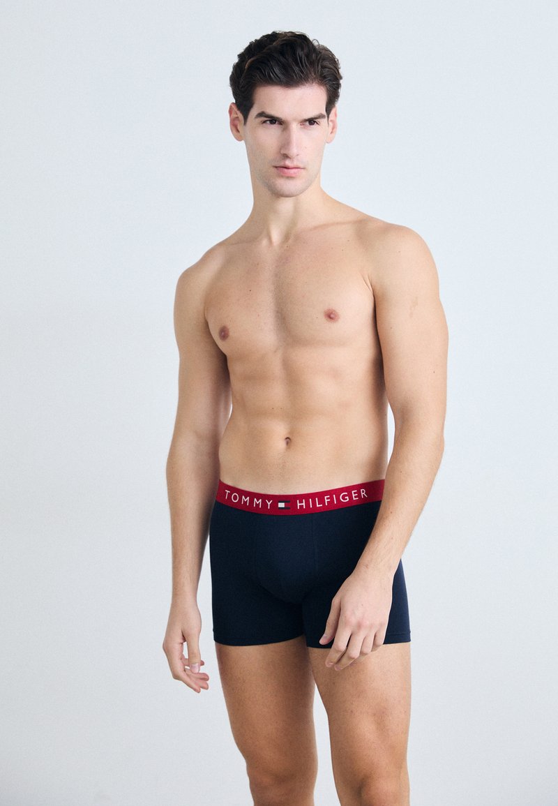 Boxeri Tommy Hilfiger în navy cu un elastic roșu ce prezintă textul logo-ului, realizați dintr-un material neted și elastic. Modelul stă într-o poziție încrezătoare.