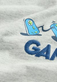 Harmaa kangaspaita, jossa on sininen brodeerattu teksti "GAME" ja värikäs grafiikka peliohjaimista, joissa on nappeja ja johtoja.