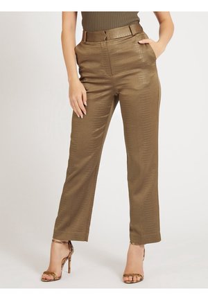 Mujer con pantalones marrones rectos con textura brillante, una mano en el bolsillo, combinados con sandalias de tacón alto con estampado de serpiente sobre fondo liso.