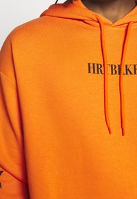 Sweat à capuche orange vif avec texte partiel noir "HRTBLKR" sur la poitrine et cordons orange assortis, porté par une personne à la peau foncée.