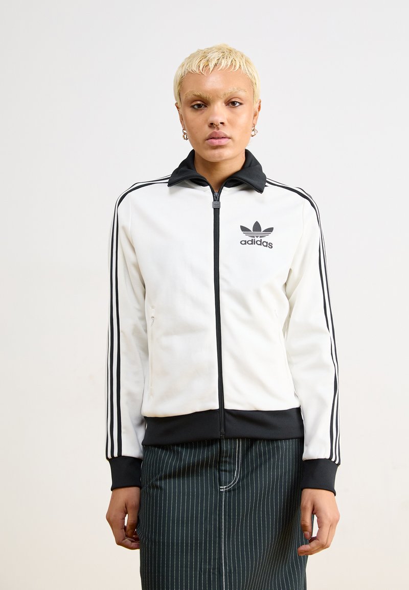 White zip-up jacket with black collar and three black stripes along sleeves. Features embroidered black Adidas logo on chest and smooth fabric.