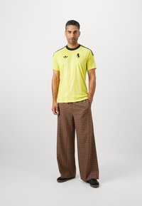 Vyras stovi vilkėdamas geltoną Adidas sportinį marškinėlį ir rudus plačius languotus kelnes su juodais batais prieš paprastą foną.