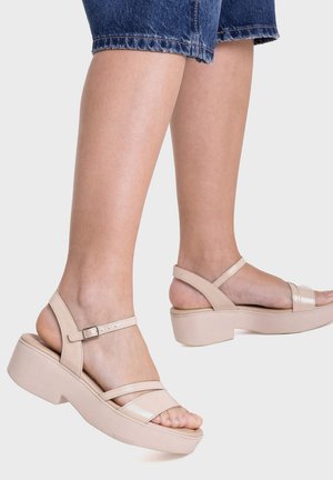 Pies con sandalias beige de plataforma con correas en el tobillo, combinadas con pantalones vaqueros azules recortados sobre un fondo liso.