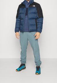 Puffer jacket in navy and black, textured fabric, paired with a blue fleece and light grey sweatpants. Sneakers feature a turquoise and black design.