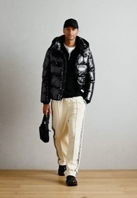 Giacca a vento nera lucida con cappuccio, felpa nera sotto, pantaloni crema con strisce laterali e sneakers nere, mentre tiene una borsa piccola.