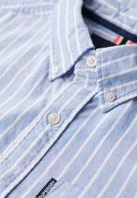 Lichtblauwe gestreepte shirt met een button-down kraag, witte knopen en een label met "Superdry." De stof heeft een gladde textuur.
