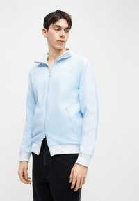 Ung mann iført lyseblå hettejakke med glidelås og hvite striper på ermene og svarte bukser, ser sidelengs mot en ensfarget bakgrunn.