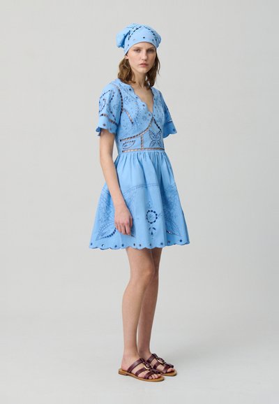 Vestido de algodón azul con mangas cortas y vaporosas, dobladillo festoneado y patrones florales bordados. Combinado con un pañuelo azul a juego y sandalias marrones.