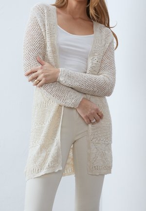 Femme portant un cardigan crème en maille ajourée sur un débardeur blanc et un pantalon crème, les mains croisées montrant des bagues en or.