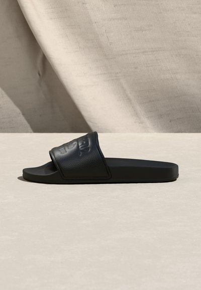 Μαύρη πλατφόρμα σανδάλι με υφασμένη λουρίδα και ανάγλυφο λογότυπο, τοποθετημένη πλαγίως πάνω σε φωτεινή επιφάνεια με απαλό ριχτό φόντο.