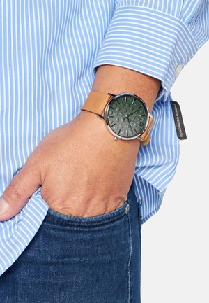 Brauner Lederarmbanduhr mit mattschwarzem Zifferblatt, das erhabene Punkte zeigt, silbernes Metallgehäuse, getragen an einem Handgelenk mit Jeans und gestreiftem Hemd.