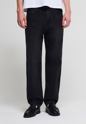 Uomo che indossa jeans neri dritti, mocassini neri in pelle e una camicia bianca in piedi davanti a uno sfondo semplice.