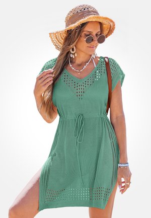 Donna che indossa un abito copricostume da spiaggia verde a maglia, cappello da sole, occhiali da sole rotondi, collana di conchiglie e braccialetti, con una borsa a tracolla marrone.
