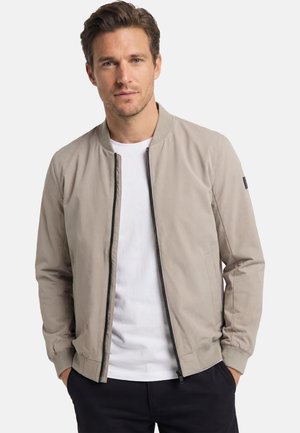 Mann mit hellbraunem Haar, trägt eine beige Reißverschlussjacke über einem weißen T-Shirt und schwarzen Hosen, steht mit den Händen in den Taschen.