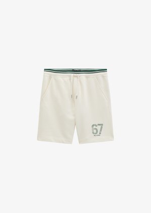 Giovane uomo che indossa una felpa con cappuccio color crema con logo e pantaloncini abbinati numerati 67, in piedi con le mani in tasca su sfondo bianco.