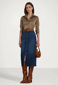 Sieviete, tērpusies brūnā satīna blūzē, augsta vidukļa džinsa midi svārkos ar priekšējo šķēlumu, brūnā ādas jostā, ceļgala augumā zābakos un nesot mazu brūnu somiņu.