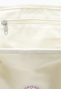 Kreminės spalvos drobės maišo vidus, su užtrauktuku kišene, struktūrizuotu dizainu ir apvaliu logotipo pachat su raudonais ir mėlynais akcentais.