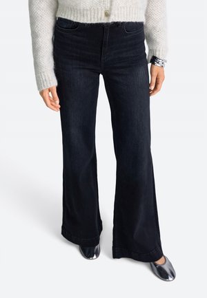 Schwarze Jeans mit weitem Bein aus Denim, mit hoher Taille und dunkler Waschung. Kombiniert mit einem leichten, strukturierten Strickpullover und metallischen Schuhen.