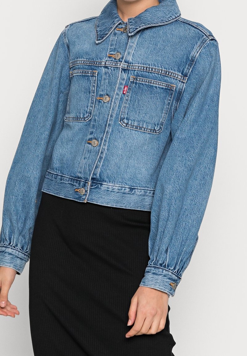 Veste en denim bleu clair avec un col, fermeture à boutons, deux poches poitrine et manches froncées. Présente des détails cousus et le logo de la marque.