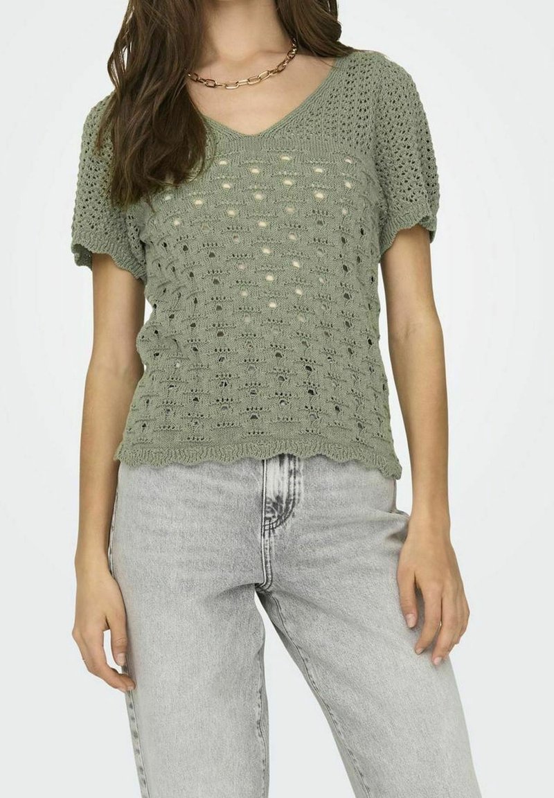 Top de punto de color verde oliva, de manga corta y con escote en V, textura de punto abierto, dobladillo festoneado y corte relajado. Llevado con jeans gris claro.