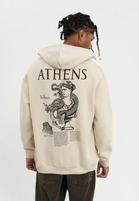 Beige hoodie with a graphic print on the back featuring a bust, serpent, and text. Ribbed cuffs and hem, soft cotton material.