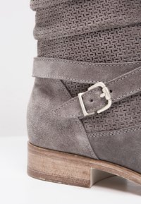 Bottine en daim gris avec tige perforée, sangle décorative, boucle argentée et talon bloc en bois sur fond blanc.