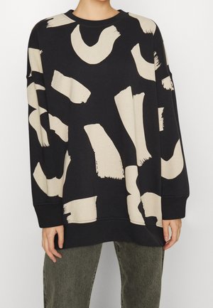 Person iført en sort oversized sweatshirt med store beige abstrakte penselstrøg og mørkegrønne bukser.