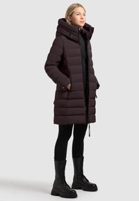 Ciemnofioletowa kurtka puchowa z wysokim kołnierzem, kieszeniami na zamek i smukłym krojem. Noszona z czarnymi legginsami i botkami w stylu wojskowym.