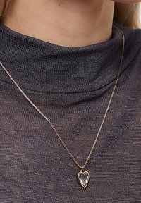 Rose gold kædedesign med et hjerteformet vedhæng, der har en transparent midte, præsenteret mod et struktureret mørkegråt stof.