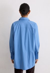 Persoon met donker haar in een knot, gekleed in een losvallende, langermige blauwe shirt, gezien van achteren tegen een effen witte muur.