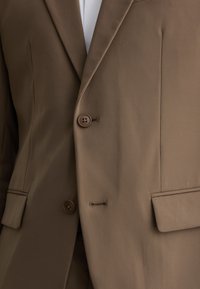 Blazer doppio petto marrone realizzato in tessuto liscio, con due bottoni, tasche laterali e un design minimalista con linee pulite.
